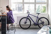 Frau Sitzt an Schreibtisch, im Hintergrund steht ein Fahrrad am Fenster.