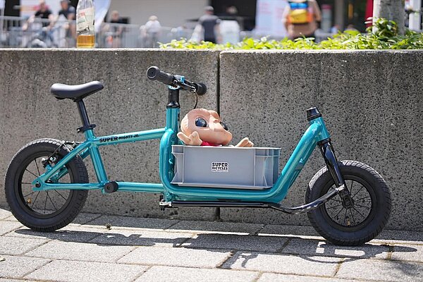 türkises Lastenlaufrad Super Mighty junior an Mauer gelehnt mit Puppe in der Kiste