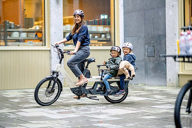 Marktübersicht E-Bikes 2024. Eine Frau fährt mit zwei Kindern auf dem Lastenrad.