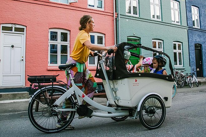 Neu sollen so viele Kinder in mit Sitzen ausgestatteten Lastenvelos transportiert werden, wie vom Hersteller vorgesehen sind. Frau mit Cargobike und Kindern vor bunter Häuserzeile