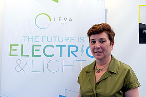 Eine Frau mit grüner Bluse steht vor einer weissen Wand.