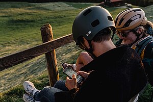 Zwei Personen mit Fahrradhelm sitzen auf einer Bank und schauen auf ein Smartphone.