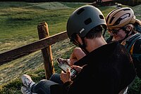 Zwei Personen mit Fahrradhelm sitzen auf einer Bank und schauen auf ein Smartphone.