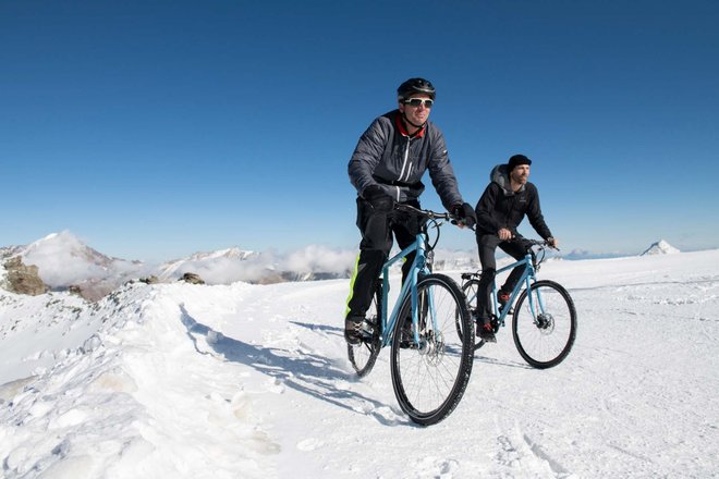 Zwei Männer fahren auf Velos durch den Schnee. Im Hintergrund sieht man Berge