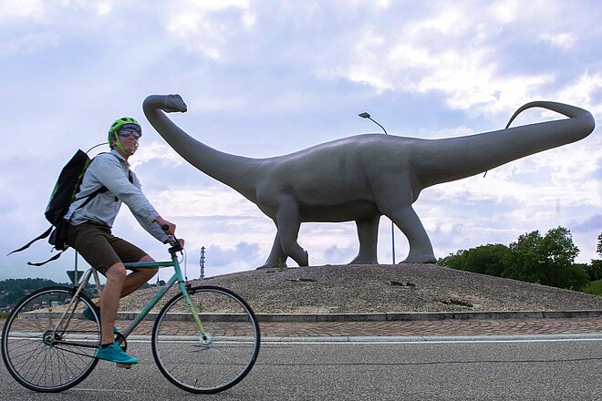 Ein Mann fährt auf dem Velo. Im Hintergrund steht eine Dinosaurier-Statue