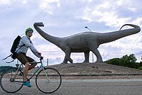 Ein Mann fährt auf dem Velo. Im Hintergrund steht eine Dinosaurier-Statue