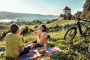 Route 1291. Die beste E-Bike Tour der Schweiz. Zwei Personen sitzen auf einer Picknickdecke und trinken Wein.