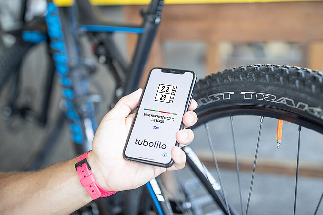 Eine Hand hält ein Smarthone, das auf dem Display Informationen zum Reifendruck des im Hintergrund sichtbaren Mountaibikes zeigt