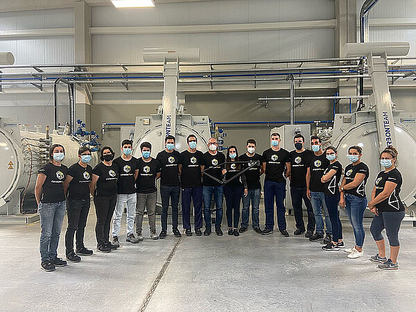 Männer und Frauen posieren mit Nasen-Mund-Schutz in einer Fabrikhalle für ein Foto.