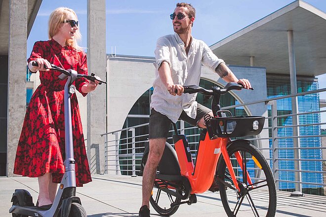 Eine Frau trägt ein rotes Kleid und steht auf einem E-Scooter, neben ihr sitzt ein Mann mit kurzen Hosen auf einem Pedelec