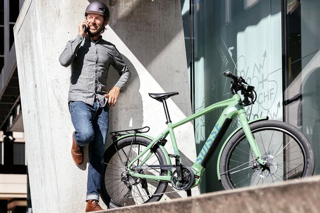Rent a Bike Tour de Suisse E-Bike Miete für Unternehmen. Ein Mann mit Fahrradhelm und grauem Hemd steht neben einem grünen E-Bike.