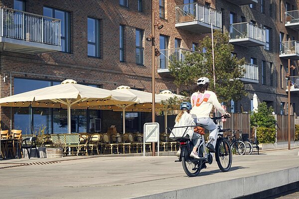 Frauen sind häufiger mit Kindern unterwegs als Männer. Frau mit Kind auf Longtail-Velo fährt entlang eines Cafes