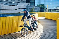 Deloitte E-Mobility Survey 2022. Ein Mann fährt mit einem E-Bike, auf dem Gepäckträger sitzen zwei Kinder in Kindersitzen. Alle tragen Fahrradhelme.