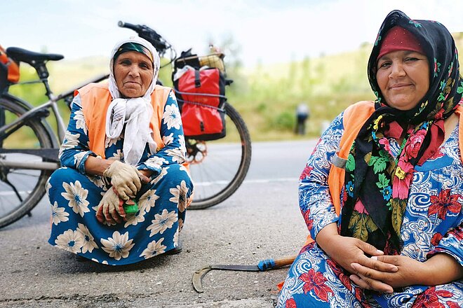 Zwei Frauen in volkstümlicher Kleidung kauern am Boden, hinter ihnen steht ein Elektrovelo