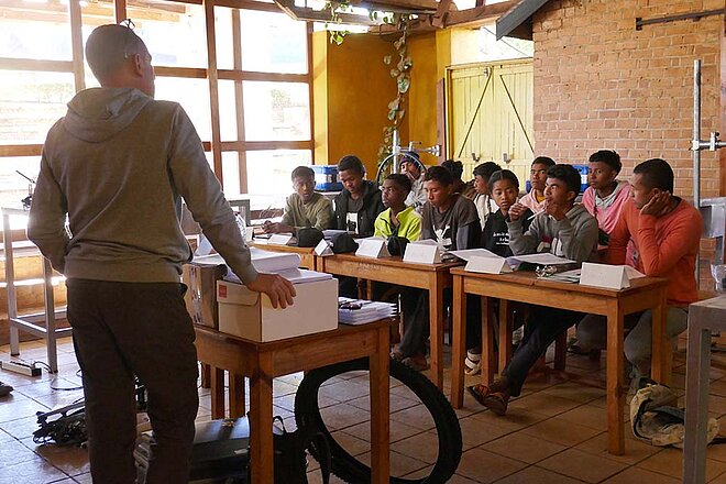 Ein Mann steht in einem Schulzimmer. An Tischen sitzen junge Menschen.