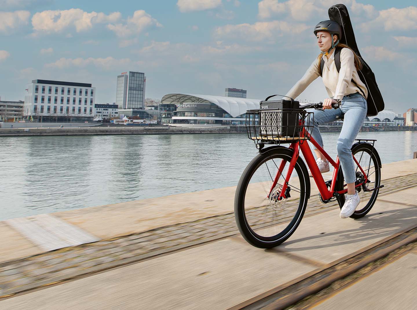 Die leichtesten E-Bikes des Jahres 2023. Light Support E-Bikes. Eine junge Frau fährt auf einem roten E-Bike. Auf dem Rücken trägt sie eine Gitarre.
